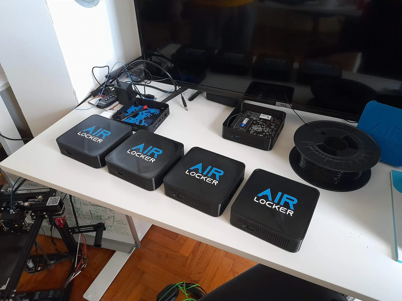 A workbench with four finished AIR LOCKER units, one opened donor mini PC showing its board, a spool of black filament and a 3D printer under the desk