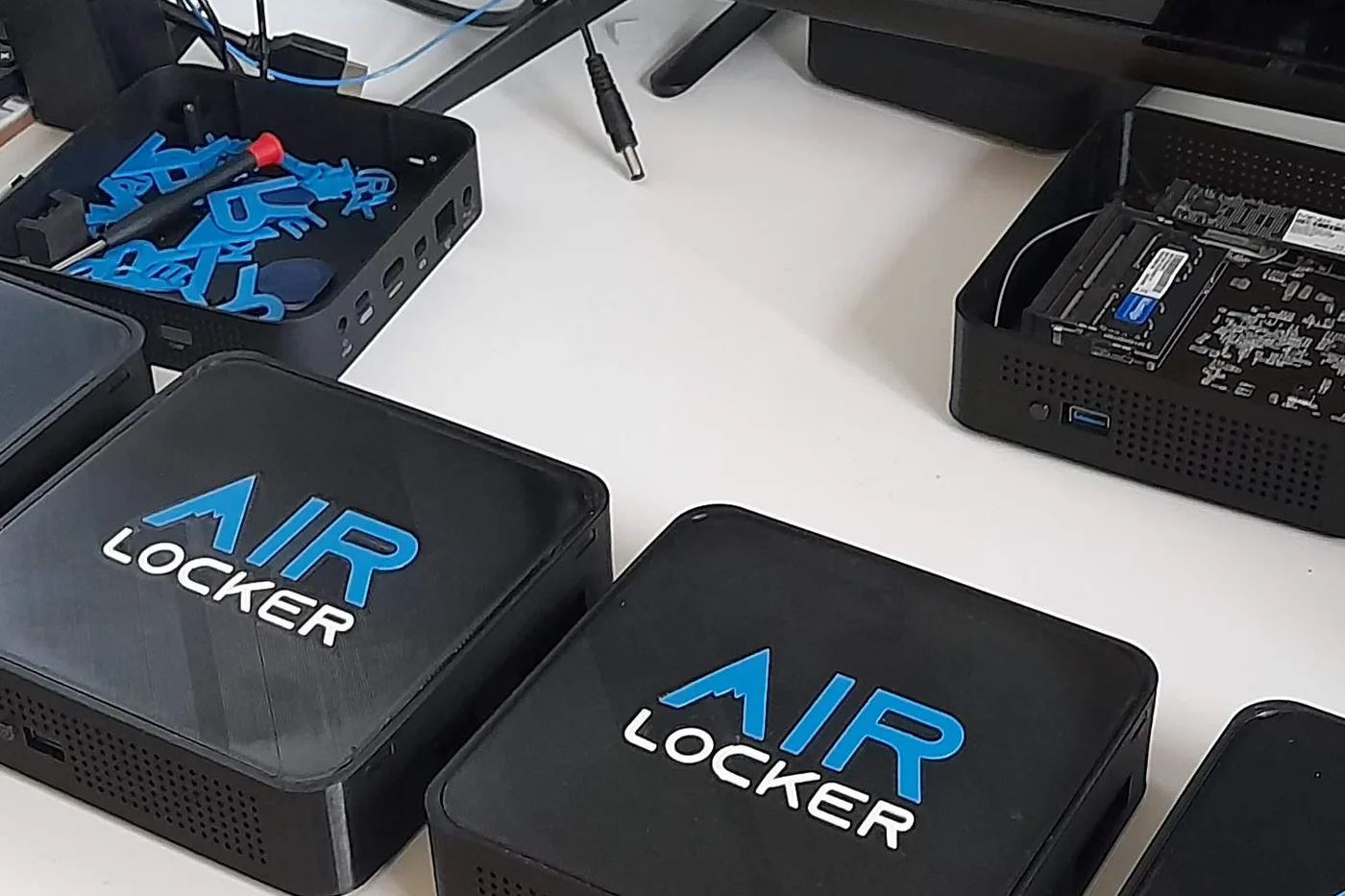 One AIR LOCKER enclosure upturned and used as a parts tray, filled with blue printed offcuts and a screwdriver, beside two finished units and an opened donor machine