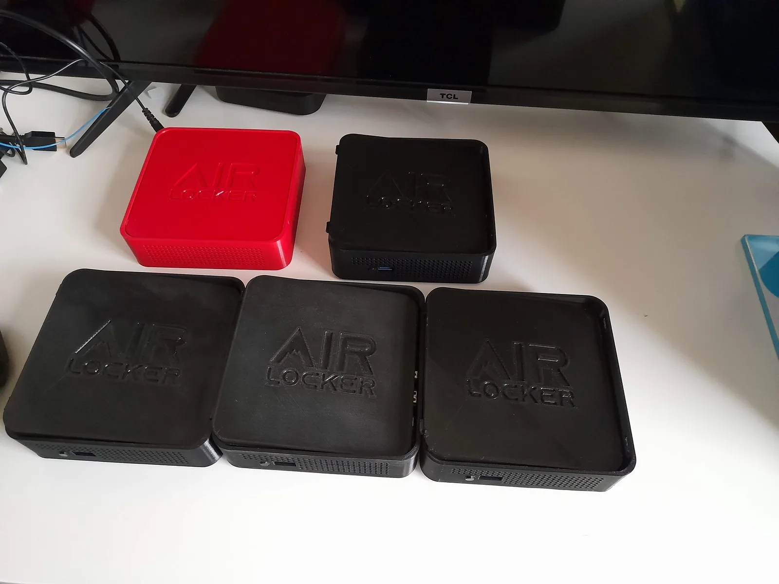 Five AIR LOCKER enclosure iterations grouped on a desk, one red and four black in different sizes, in front of a wall-mounted TV