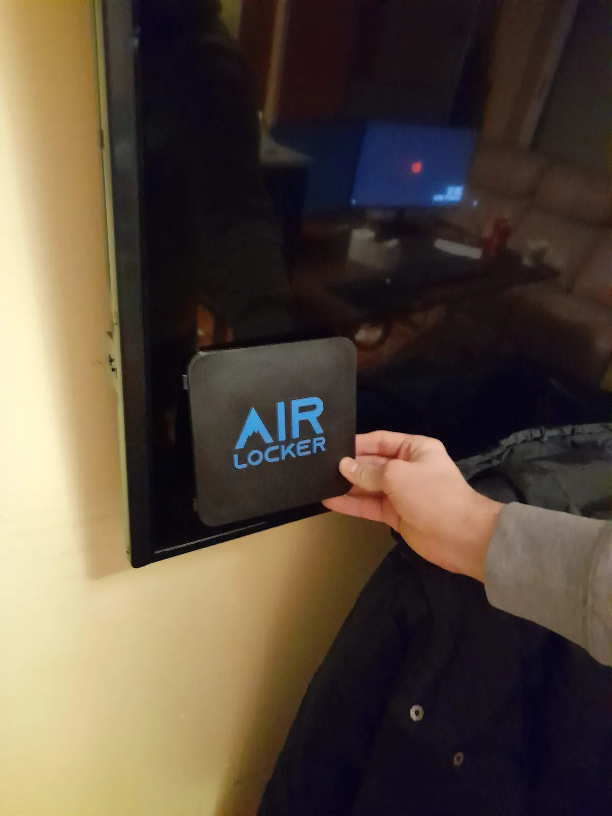 A hand holding an AIR LOCKER unit against the back of a wall-mounted television to check clearance