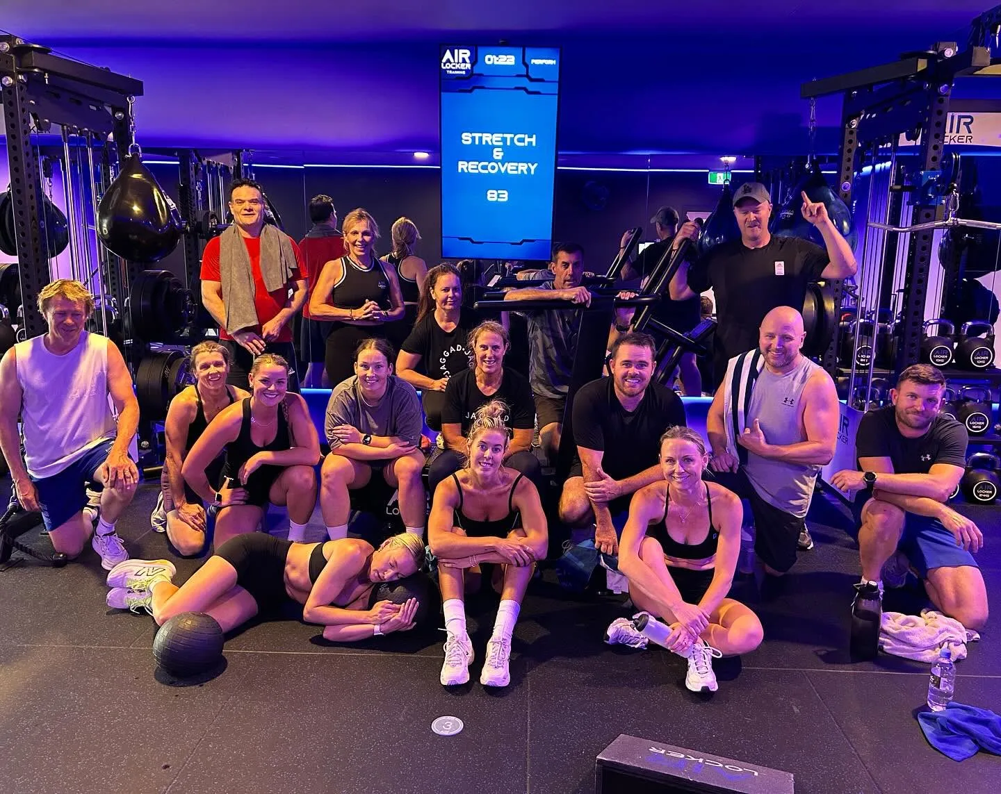 An AirLocker studio class gathered in front of a wall-mounted screen showing STRETCH & RECOVERY with a live class timer